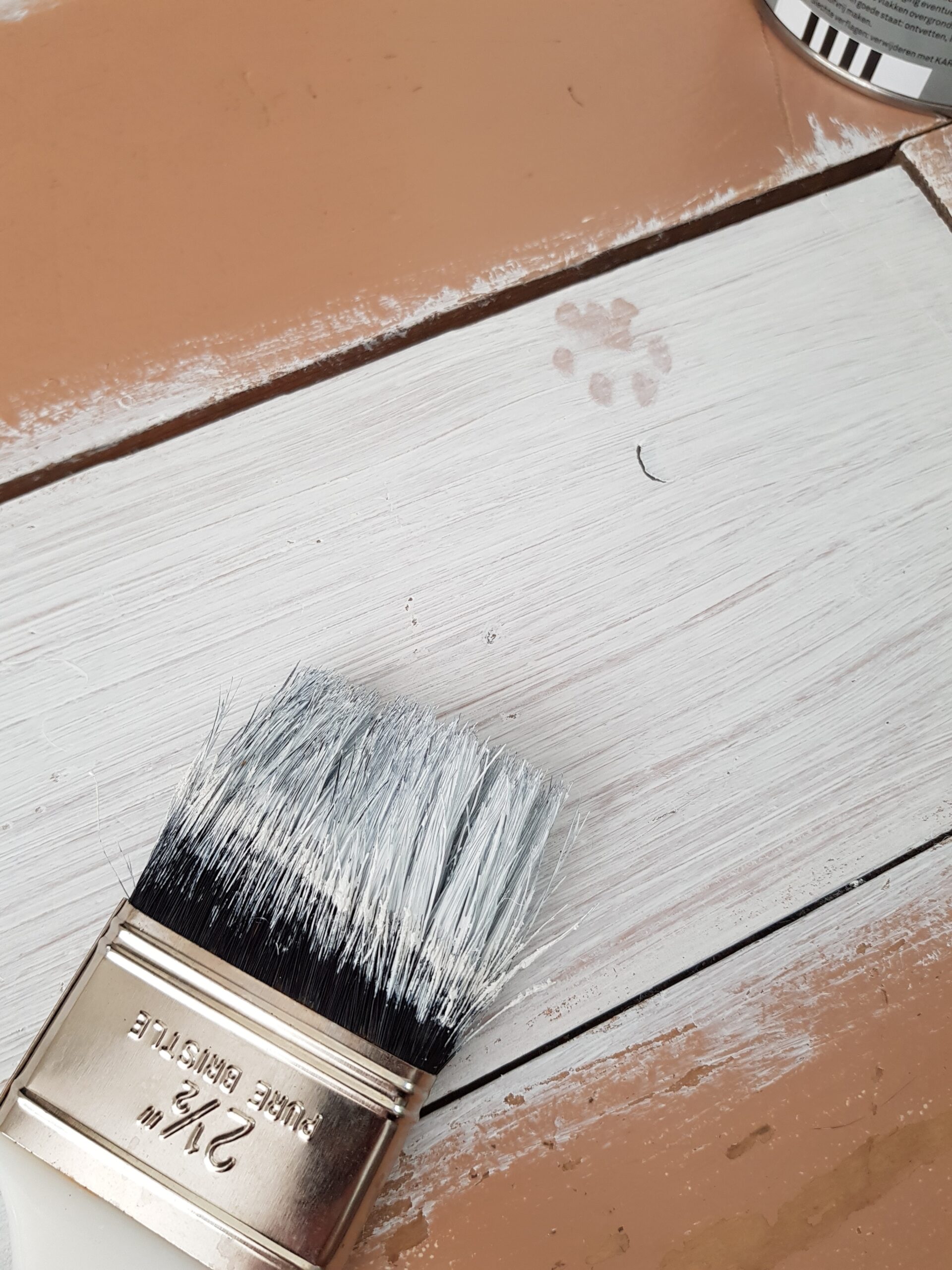 A paintbrush applying white paint onto a wooden surface.