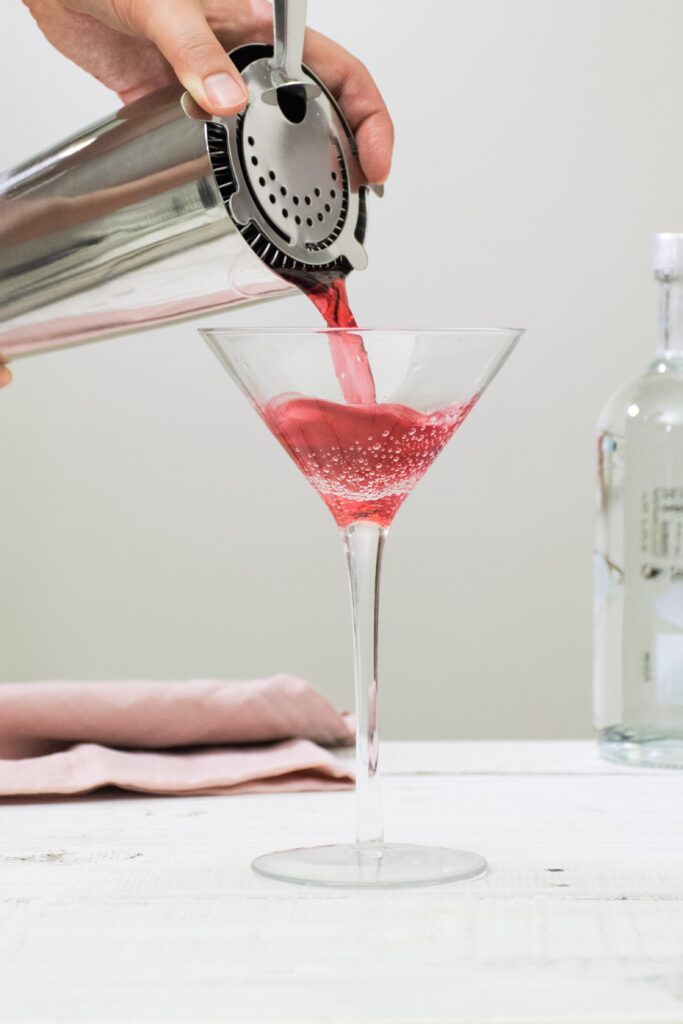 A person pouring a cosmopolitan cocktail into a martini glass