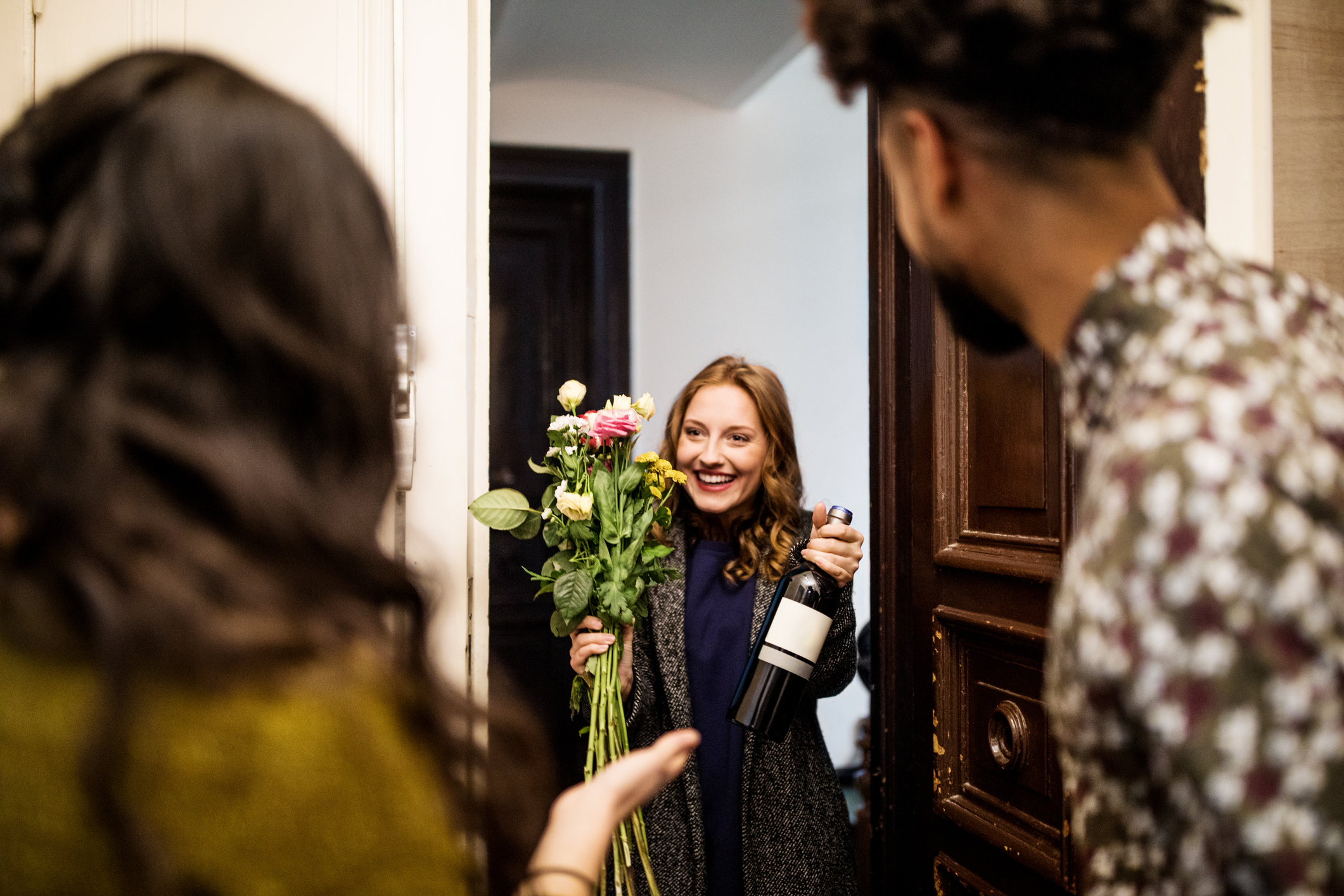 Young woman holding bouquet and wine bottle while visiting friends for dinner party