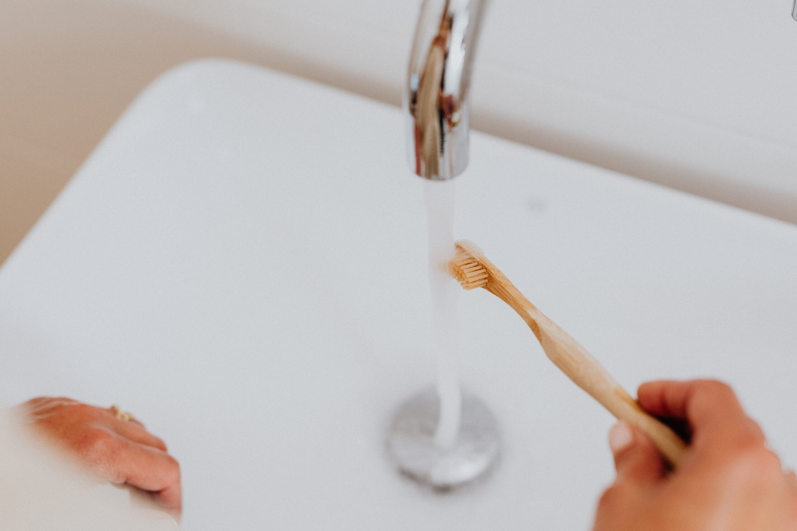 A hand holding a wooden toothbrush under a running faucet
