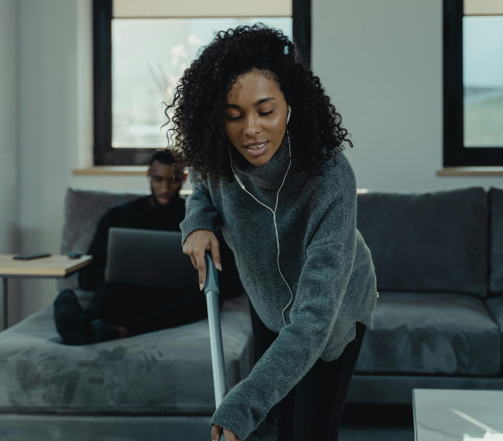 Woman in gray shirt holds vacuum handle while man in background sits on couch
