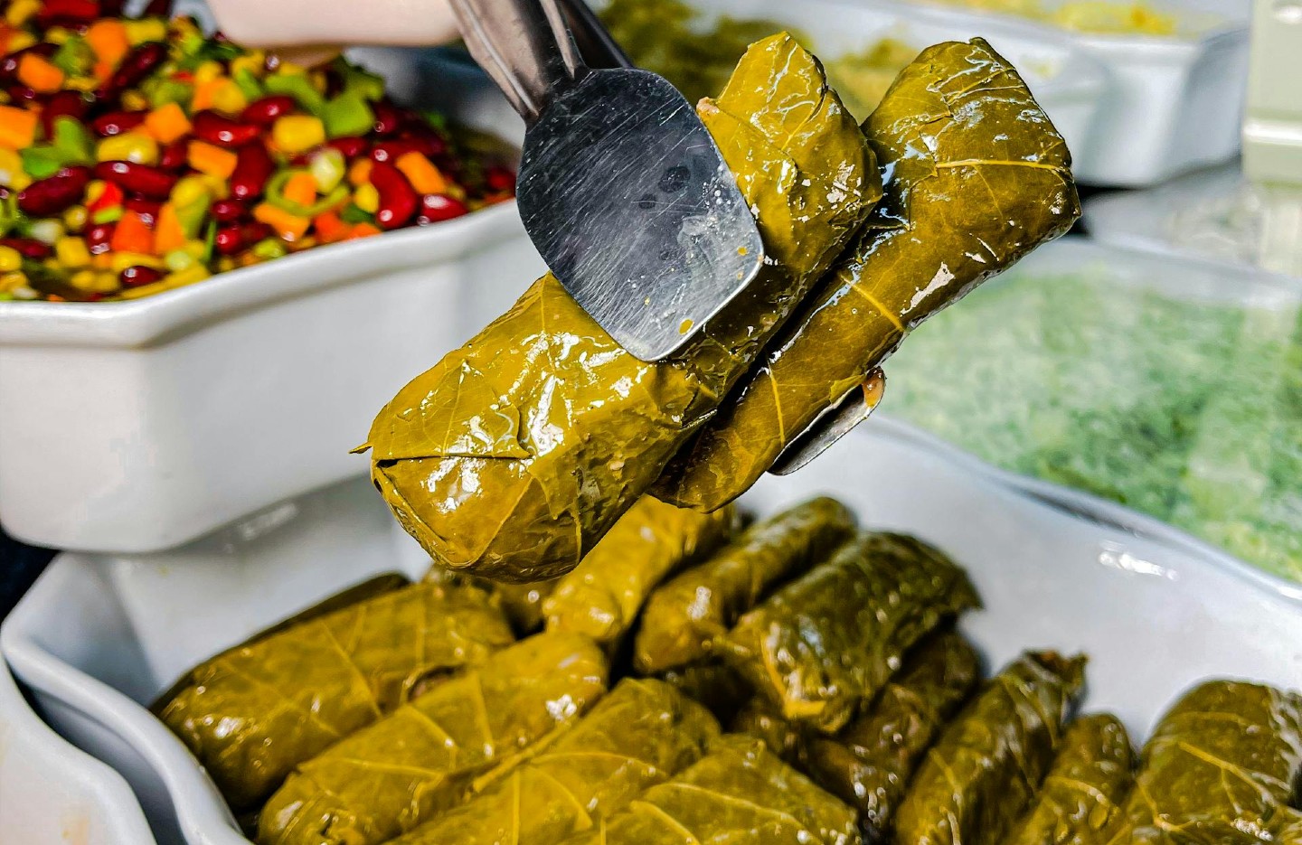Tongs holding stuffed grape leaves with mixed vegetables in a bowl in the background